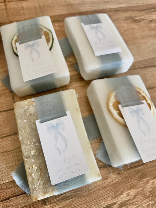 May include: Four white soap bars wrapped in blue ribbon with a thank you tag attached to each. The tags have a watercolor bow design and text that reads &#39;Thank you for helping us tie the knot&#39; and &#39;Thank you Mrs. Brooks EST. 2021&#39;. The soap bars are decorated with dried citrus slices.
